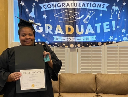  New dental assistant graduate at Tulsa DAS smiling with her program certificate in front of the graduation banner. 