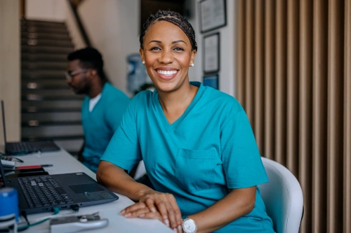 Female medical assistant smiling and using laptop alongside other medical assistant on computer